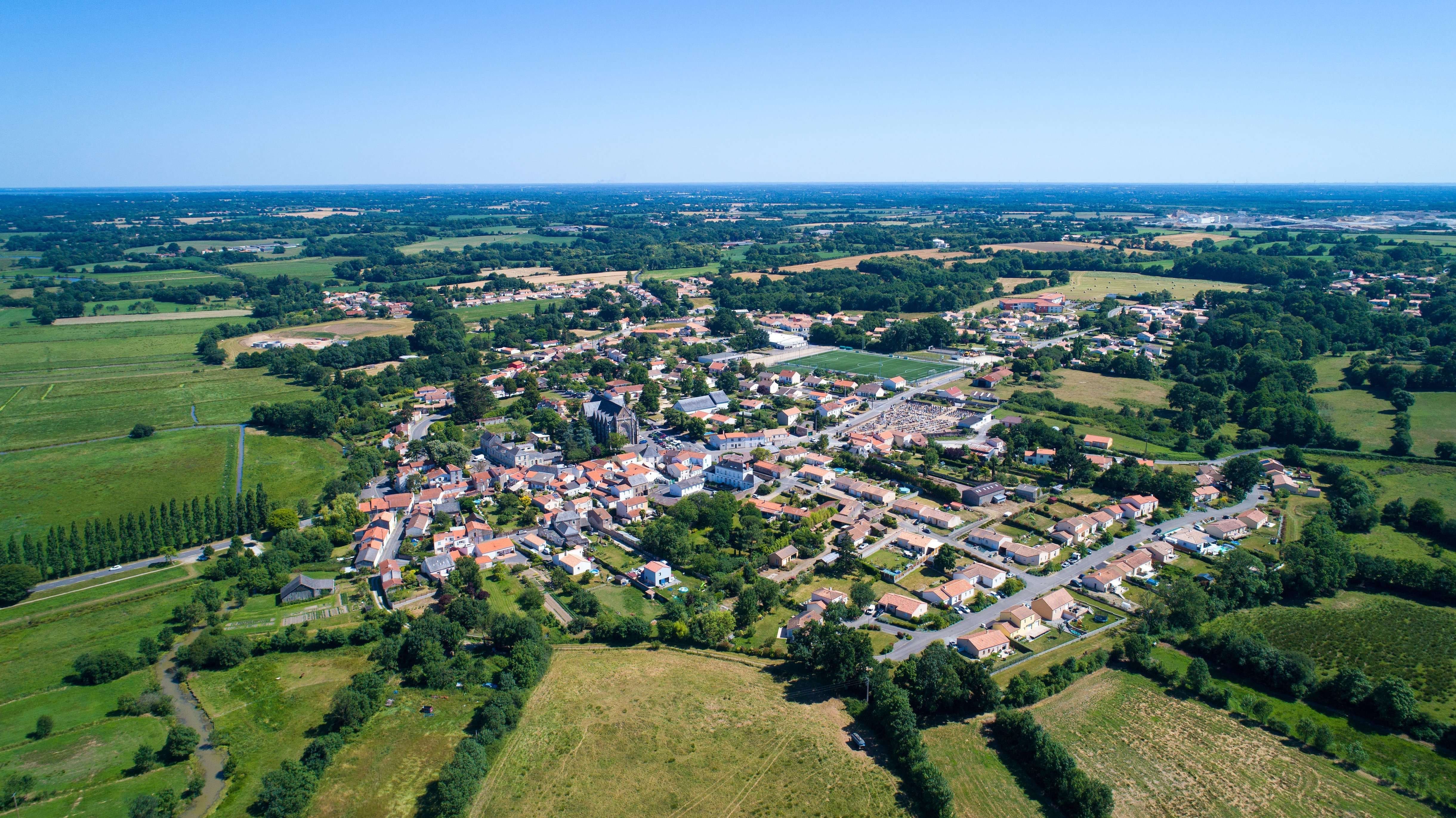 Luftaufnahme einer ländlichen Ortschaft, umgeben von Feldern, Wiesen und weitläufiger Landschaft.