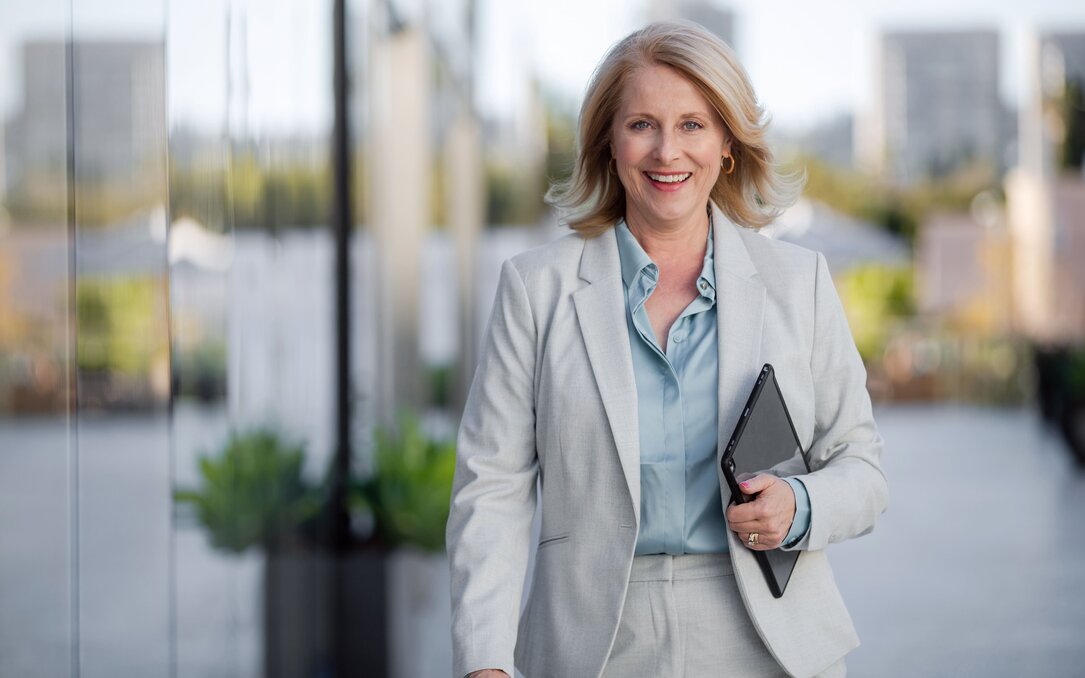 Geschäftsfrau geht lächelnd im Außenbereich eines Bürokomplexes und hält ein Tablet in der Hand.
