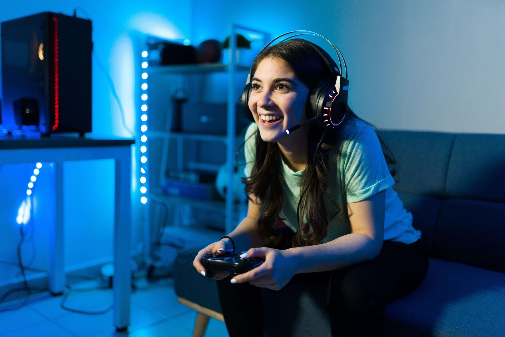 Junge Frau spielt mit einem Controller und Headset begeistert ein Videospiel in einem blau beleuchteten Raum.